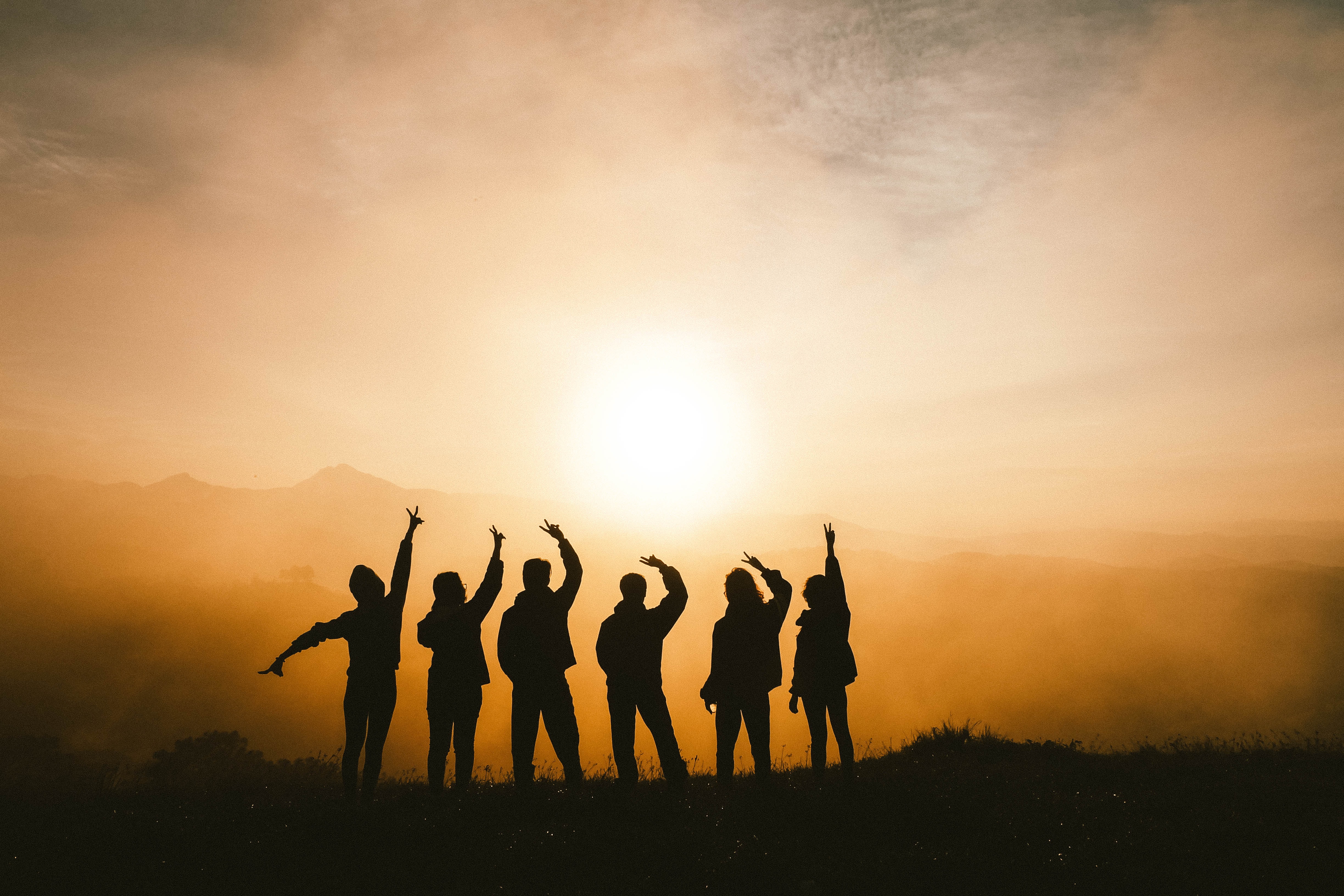 six people on a mountain in front of a sunset raising their hands in a peace sign